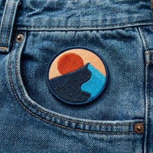 May include: Embroidered patch featuring a mountain scene with a setting sun. The patch has a circular shape with a tan background, a red sun, and a mountain in shades of blue and turquoise. The patch is sewn onto a blue denim pocket.