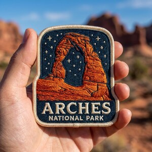 May include: A rectangular embroidered patch with rounded top corners featuring a dark blue night sky filled with white stars above a rust-orange depiction of Delicate Arch. The text ARCHES NATIONAL PARK is embroidered in white at the bottom.