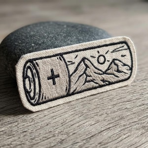 May include: Embroidered patch featuring a battery design with a mountain landscape and sun. The patch has a beige background with black stitching. The battery has a plus sign. Ideal for jackets, bags, or other fabric items.