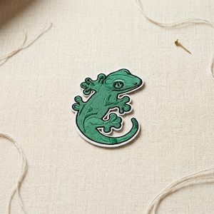May include: A green embroidered gecko patch with a white border. The gecko has detailed stitching and a curled tail. The patch is on a beige fabric background with a few strands of thread and a small pin.