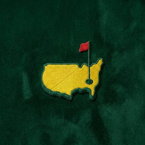 May include: Embroidered patch featuring the outline of a yellow map with a red flag on a green background. The patch has a green border and is likely a logo or emblem.