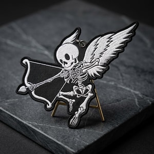 May include: Embroidered patch featuring a skeleton with angel wings, holding a bow and arrow. The patch is black and white, with detailed stitching. The design is a unique take on a classic theme.