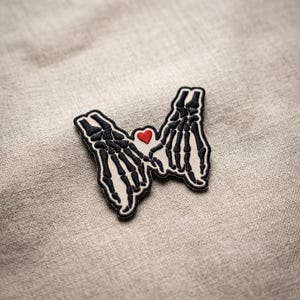 May include: Embroidered patch featuring two black skeleton hands holding a red heart. The patch has a white background with a black border. The design is on a beige fabric surface.