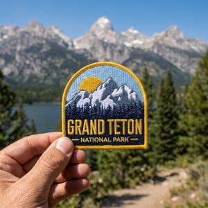 May include: A semi-circular embroidered patch featuring a mountain range with snow-capped peaks, a yellow sun, and a dark blue forest. The text GRAND TETON NATIONAL PARK is stitched in yellow capital letters against a dark blue background.