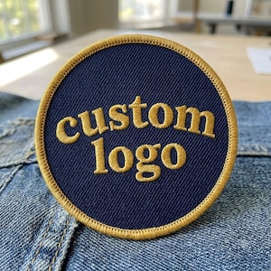 May include: A circular embroidered patch with a dark navy blue background and a gold border. The text custom logo is stitched in gold thread across the center of the patch in a rounded, lowercase font. The patch is placed on blue denim fabric.