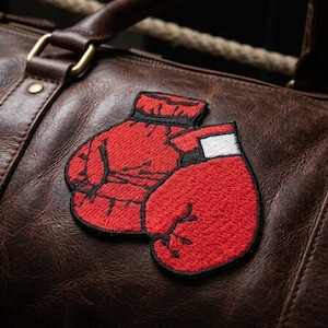 May include: A close-up of a brown leather duffel bag with a pair of red boxing glove patches. The gloves are embroidered with black outlines and white accents. The bag has a handle and gold-colored hardware.