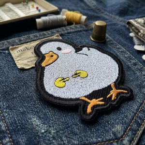 May include: Embroidered patch featuring a cartoon duck with a light blue body, black outline, and yellow feet and beak. The patch is on a denim background, with sewing supplies like needles, thread, and a thimble visible.