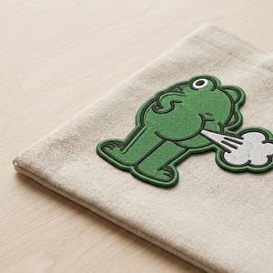 May include: A beige tote bag with a green frog patch. The frog is depicted from the rear, with a cloud of white smoke emanating from its backside. The patch has a black outline.