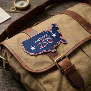 May include: Canvas messenger bag with brown leather accents and straps. A blue, white, and red patch in the shape of the United States is affixed to the front. The patch reads "AMERICA 250" with two white stars.
