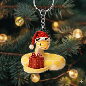 May include: A yellow snake-shaped Christmas ornament wearing a red Santa hat with the name "CHERRY" on it. The snake is holding a red gift box with a red bow. The ornament hangs from a silver chain, set against a blurred Christmas tree background.