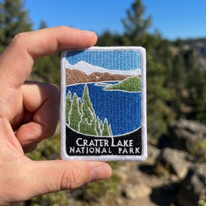 May include: A rectangular embroidered patch featuring a landscape scene of Crater Lake National Park. The design includes a blue lake, green pine trees, brown mountains, and a light blue sky. The text Crater Lake National Park is in white.