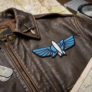 May include: A close-up of a brown leather pilot jacket with a blue and white embroidered patch. The jacket has a brass propeller pin on the collar and a set of dog tags. A map and navigation tools are in the background.
