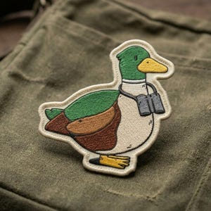 May include: Embroidered patch featuring a cartoon duck wearing binoculars. The duck has a green head, brown and green wings, and a cream-colored body. The patch is outlined in white and set against a green fabric background.