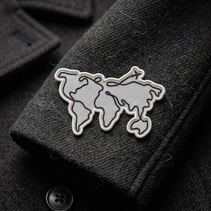 May include: A gray and white world map patch is sewn onto a dark gray wool coat sleeve. The patch features a continuous line design with a white border. A black button is visible at the bottom of the image.