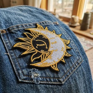 May include: Embroidered patch featuring a celestial design of a sun and moon. The patch is stitched with gold, black, and white thread and is affixed to a denim pocket. The denim has a distressed appearance.