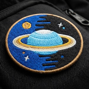 May include: Embroidered patch featuring a blue planet with rings, a yellow moon, and stars against a blue and black background. The patch is circular with a tan border and is sewn onto a black fabric.