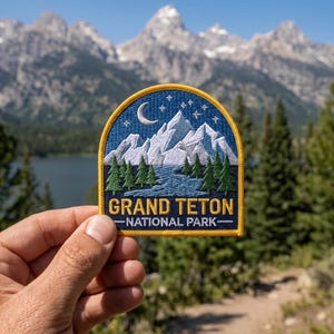 May include: A semi-circular embroidered patch featuring a night scene of the Grand Teton National Park. The design includes white snow-capped mountains, a blue lake, green pine trees, a crescent moon, and stars, with the text GRAND TETON NATIONAL PARK.
