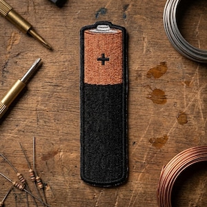 May include: An embroidered patch shaped like a cylindrical battery. The top section is copper-colored with a white terminal and a black plus sign symbol. The bottom section is solid black. The patch has a thick black border and a textured finish.