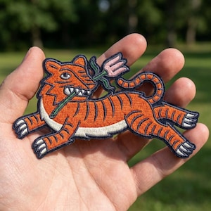May include: An embroidered patch featuring an orange tiger with black stripes and a white underbelly. The tiger is depicted in a running pose, holding a pink flower with a green stem in its mouth. The patch has a dark blue border.