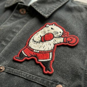 May include: Embroidered patch featuring a boxing groundhog in red shorts and boxing gloves. The patch is white, beige, and red, with black accents. The patch is sewn onto a dark gray denim jacket.