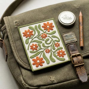 May include: Embroidered patch with orange flowers and green leaves on a white background. The patch is on a green canvas bag, next to a metal lid with the word "Lens", a pencil, and a leather strap.