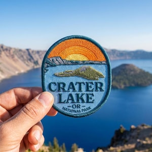 May include: A circular embroidered patch featuring a landscape of Crater Lake National Park. The design includes a bright orange and yellow sun over a blue lake and a green island, with the text CRATER LAKE NATIONAL PARK in dark blue.