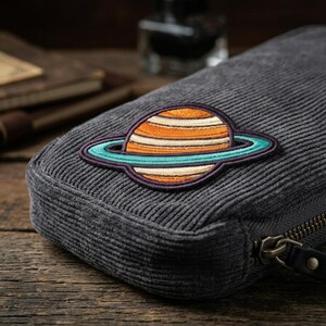 May include: A gray corduroy pencil case with a colorful embroidered planet patch. The patch features orange, white, and blue stripes with a teal ring. The case has a zipper closure and a black leather pull.