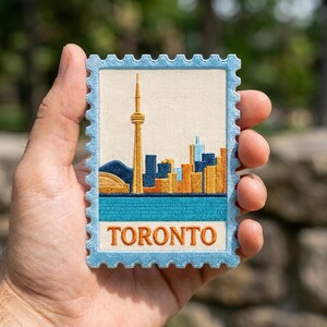 May include: A rectangular embroidered patch shaped like a postage stamp featuring the Toronto skyline. The design includes the CN Tower in gold, various buildings in blue and orange, and the word TORONTO in orange text on a cream background.