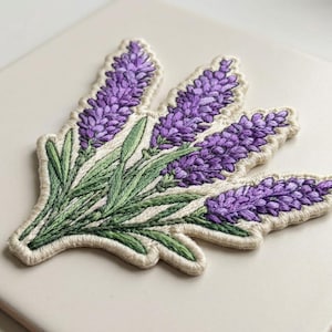 May include: Embroidered lavender patch featuring purple flower heads and green leaves. The patch has a cream-colored border and is likely for decorative purposes, such as on clothing or accessories. The design is detailed and realistic.