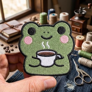 May include: An embroidered patch shaped like a green frog with black eyes, pink cheeks, and a smiling mouth. The frog is holding a white coffee mug with brown liquid and white steam rising from it, outlined in black thread on a green background.