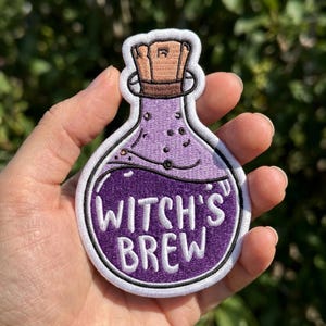 May include: An embroidered patch shaped like a potion bottle with a light purple top and a dark purple bottom. The bottle features a tan cork stopper and the white text WITCH'S BREW embroidered across the center of the dark purple liquid section.