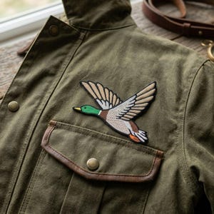 May include: An olive green jacket with a duck patch. The patch shows a flying duck with brown, beige, green, and orange embroidery. The jacket has a pocket with a brown leather trim and button closures. A compass and leather strap are visible.