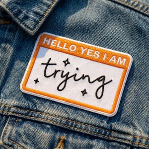 May include: A rectangular embroidered patch with a white center and an orange border. The top section features the white text HELLO YES I AM on an orange background. The center displays the black cursive word trying with four small black stars.