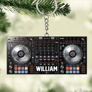 May include: A black DJ controller keychain with the name "WILLIAM" in white. The keychain features two silver-colored turntables and colorful buttons. The keychain is hanging from a silver chain and ring.