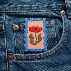 May include: Embroidered patch featuring a red poppy flower with green leaves on a pink background, framed by a blue scalloped border. The patch is affixed to the pocket of a pair of blue denim jeans.