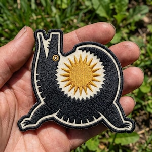 May include: An embroidered patch featuring a stylized black creature with a long snout and clawed limbs. The creature's body is circular and contains a central golden yellow sunburst design with radiating rays against a cream-colored background.