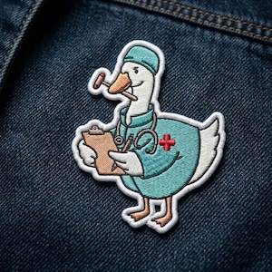 May include: Embroidered patch featuring a cartoon goose dressed as a doctor, wearing a teal uniform, stethoscope, and cap. The goose holds a clipboard and a medical hammer. The patch has a white border and is set against a dark blue denim background.