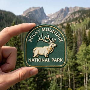 May include: A dark green embroidered patch featuring a cream-colored elk with large antlers in the center. The text "ROCKY MOUNTAIN" is arched above the elk, and "NATIONAL PARK" is printed below in cream-colored lettering with a gold border.