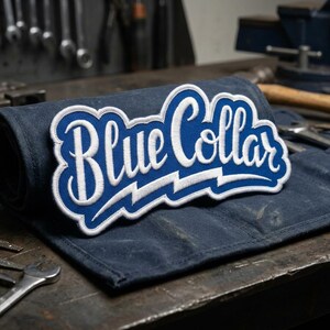 May include: A rolled-up, dark blue canvas tool roll with a large, embroidered patch. The patch features the words "Blue Collar" in white script with a blue outline and a white lightning bolt design.