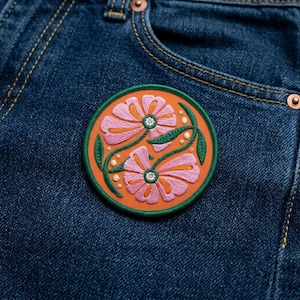 May include: Embroidered patch featuring two pink and orange flowers with green leaves and white accents, set against an orange background. The patch has a green border and is affixed to a blue denim fabric.