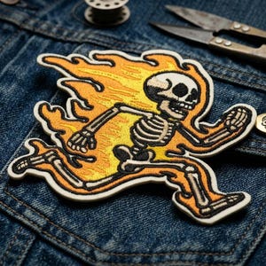 May include: Embroidered patch featuring a running skeleton engulfed in flames. The design uses black, yellow, and orange thread on a white background. The patch is placed on a denim jacket, with a skull button and sewing tools visible.