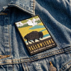 May include: A rectangular embroidered patch featuring a black bison silhouette against a landscape of yellow fields, blue water, and white geysers. The text YELLOWSTONE NATIONAL PARK is stitched in yellow and white on a black background.