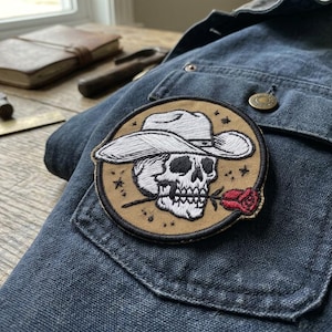 May include: Embroidered patch featuring a skull wearing a cowboy hat and holding a red rose in its teeth. The patch is circular with a tan background and black border, sewn onto a blue denim jacket.