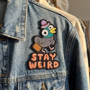 May include: A fabric patch attached to a blue denim jacket featuring a cartoon pigeon wearing a purple hat and carrying a brown briefcase. The pigeon has a teal head, gray body, and yellow beak. Below the bird, the text STAY WEIRD is in orange.