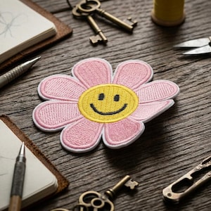 May include: A pink daisy patch with a yellow smiley face center, surrounded by craft supplies on a wooden surface. The patch is embroidered with a white border. Other items include keys, scissors, and a notebook.