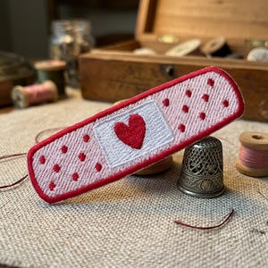 May include: Embroidered bandage patch with a red heart on a white square, surrounded by red dots. The patch is pink with a red border. A thimble, spools of thread, and a wooden box are in the background.