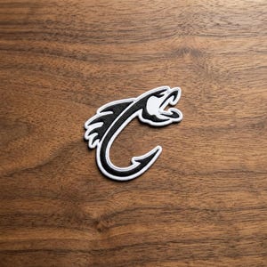 May include: A black and white embroidered patch featuring a stylized fish hook design. The hook is shaped to resemble a fish with an open mouth and fins, outlined in white thread with a solid black interior, creating a sharp graphic contrast.