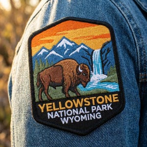 May include: A hexagonal embroidered patch featuring a brown bison standing before a waterfall, blue mountains, and an orange sky. The black border contains the yellow and white text: YELLOWSTONE NATIONAL PARK WYOMING. The patch is attached to denim.