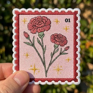 May include: A rectangular embroidered patch shaped like a postage stamp with a scalloped white border and a dark red frame. The light pink center features two dark pink carnations with green stems and yellow star icons, with the text 01.