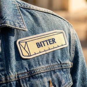 May include: A rectangular embroidered patch attached to a blue denim jacket. The patch features a light yellow background with a dark blue border and the word BITTER in dark blue capital letters, alongside a graphic of a ruler and a triangle.